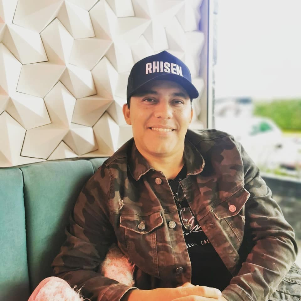 Hombre sonriente con chaqueta de camuflaje y gorra RHISEN sentado frente a una pared geométrica.